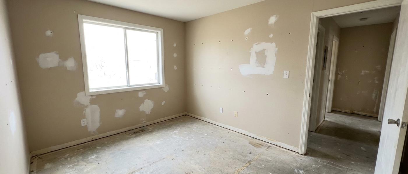 An empty rental apartment room between tenants with beige walls, several white patched spots of joint compound drying, a bare floor and an open door to a hallway